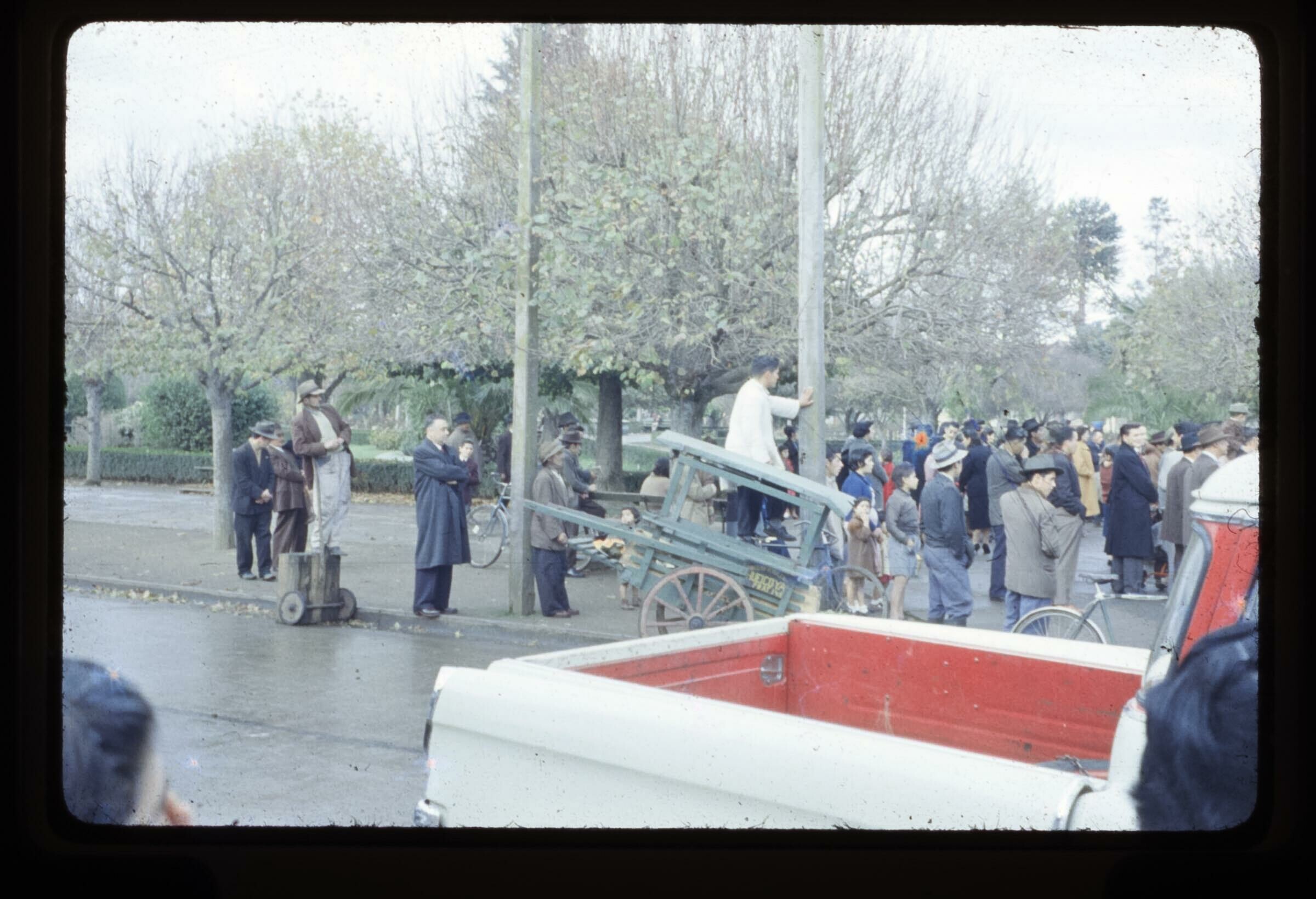 Plano entero de un grupo de personas de pie, uno de ellos arriba de un carretón afirmándose de un poste, para observar un evento en Plaza de la Concordia de La Unión