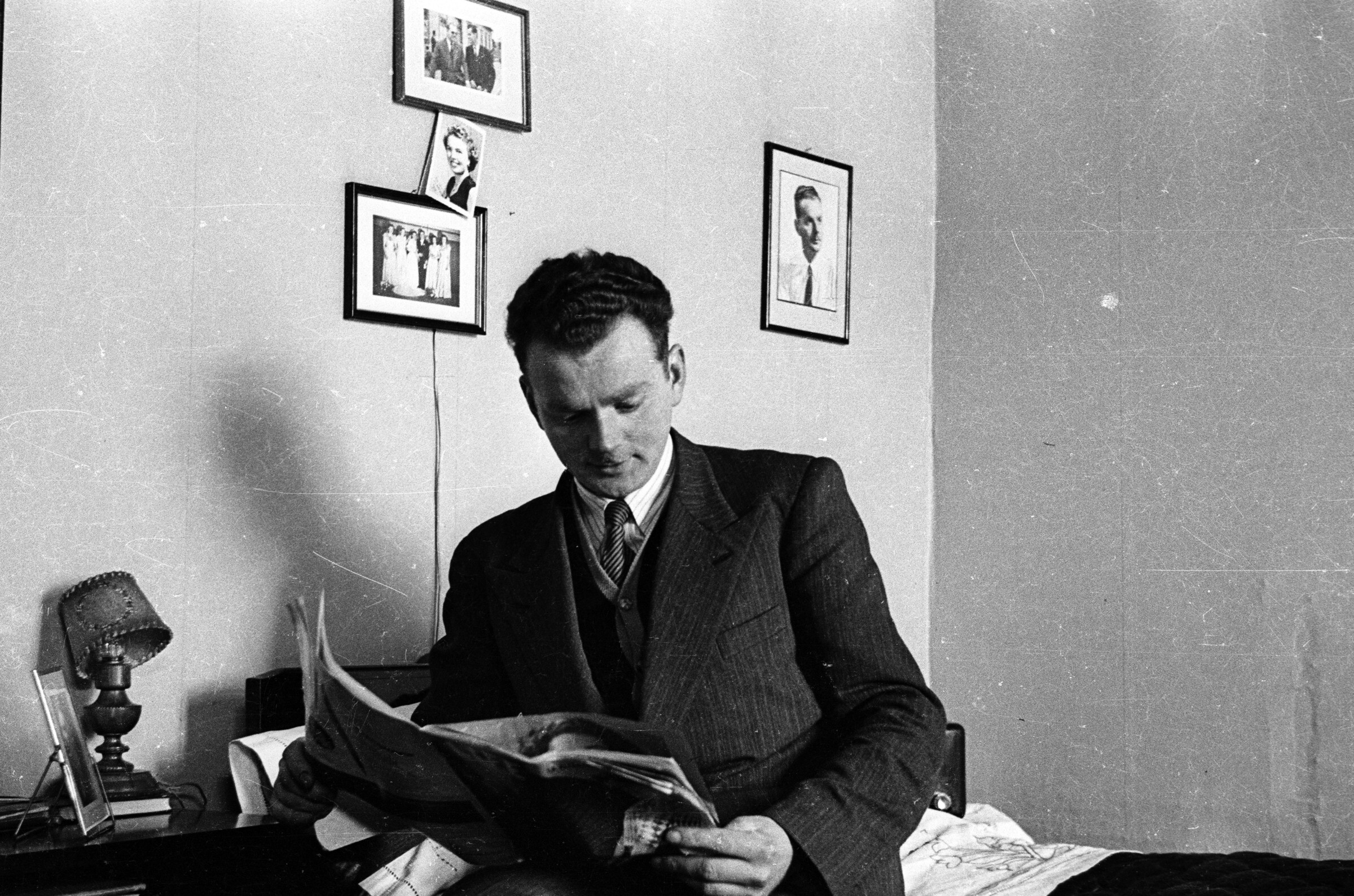 -Plano entero de Gonzalo carrasco, sentado al borde de una cama leyendo una revista llamada --Life-- con las piernas cruzadas. Se encuentra en el interior del dormitorio de una casa-