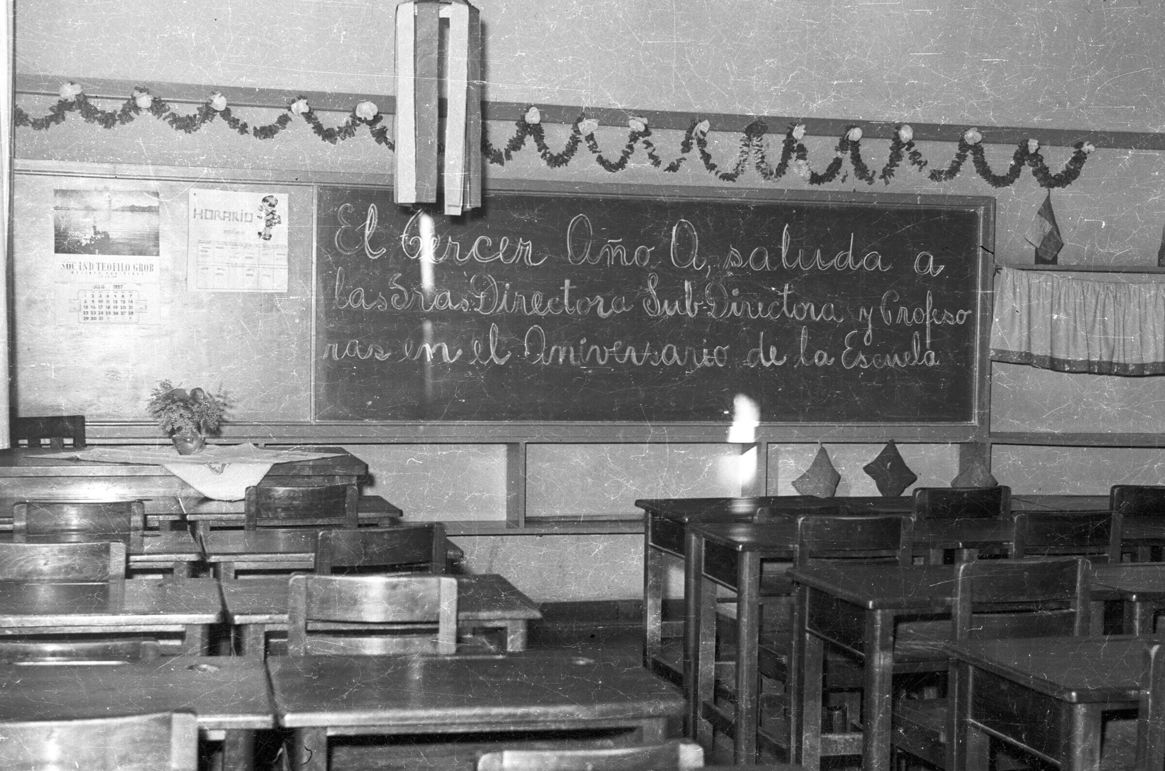 Plano entero de sala de clases con pupitres y pizarrón con un mensaje por el 54 aniversario de Escuela superior de niñas n°2