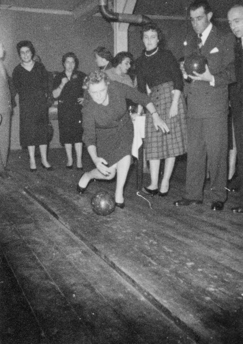 Plano entero de Olga Boettcher lanzando una bola sobre un piso de madera mientras juega bowling o también conocido como bolo americano. A su alrededor, Hombres y Mujeres le acompañan en el interior de un recinto no identificado