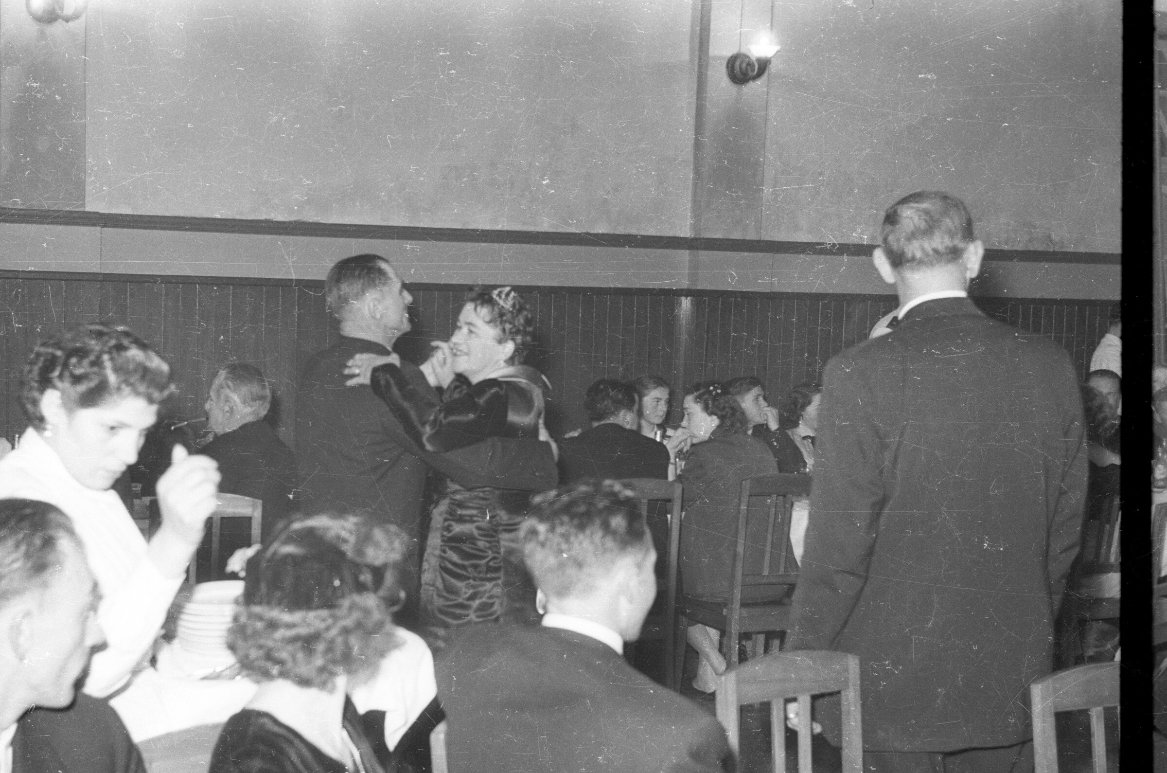Retrato de plano entero de Hombre con Traje y Mujer con Vestido bailando y sonriendo, alrededor se ven personas sentadas