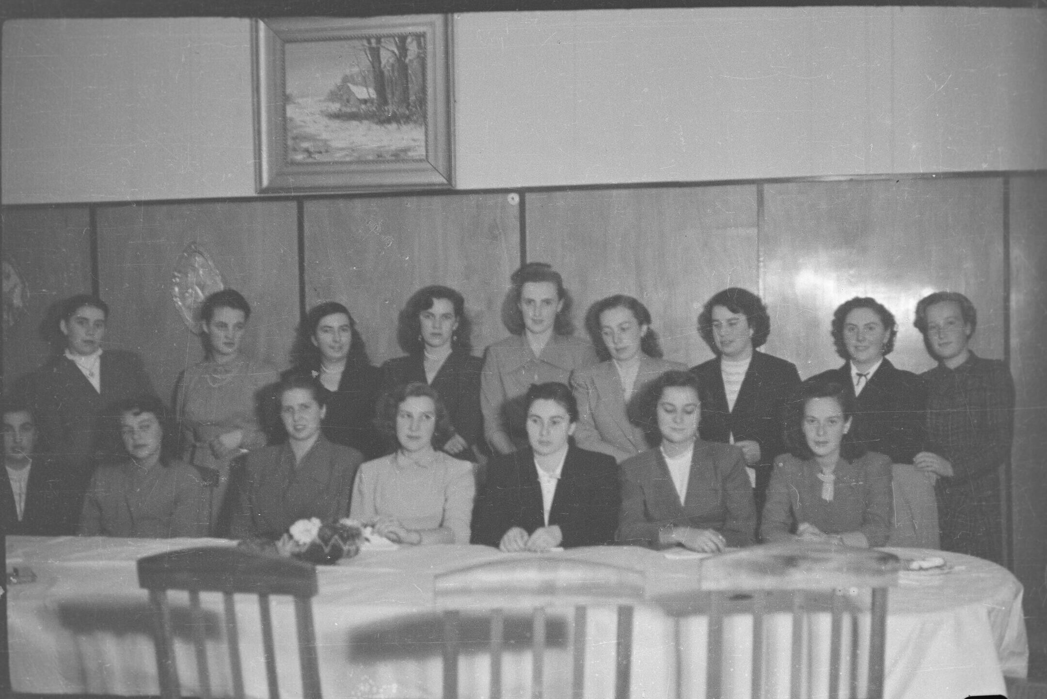 Retrato grupal de plano entero de 16 mujeres posando junto a una larga mesa en un lugar no identificado