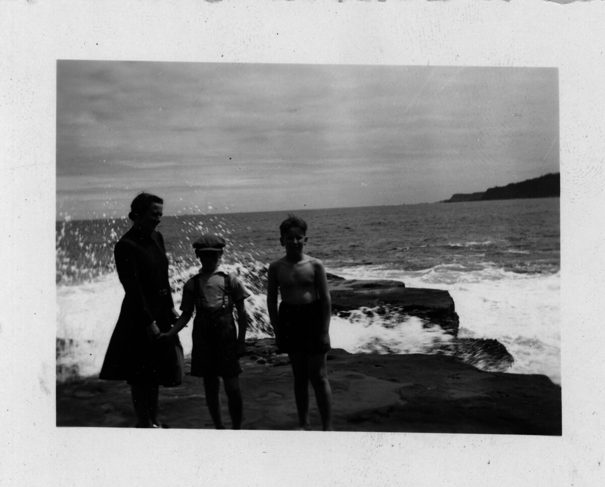 Retrato de plano entero de dos niños posando parados sobre una roca. El niño de la izquierda está tomado de la mano de una mujer. A sus espaldas, una ola rompe sobre el roquerío. La fotografía fue tomada durante un paseo a una playa no identificada