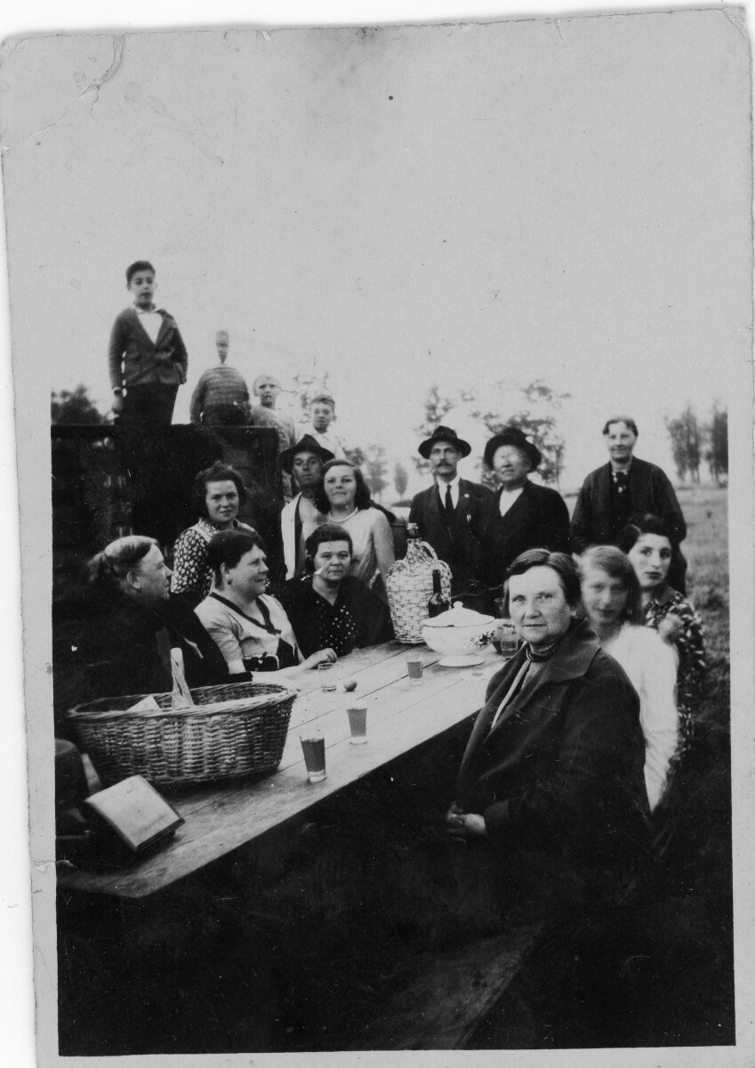 Retrato grupal de plano entero de Niños, Mujeres y Hombres posando alrededor de una mesa sobre la cual hay vasos servidos con chicha. Están compartiendo al aire libre en un lugar no identificado