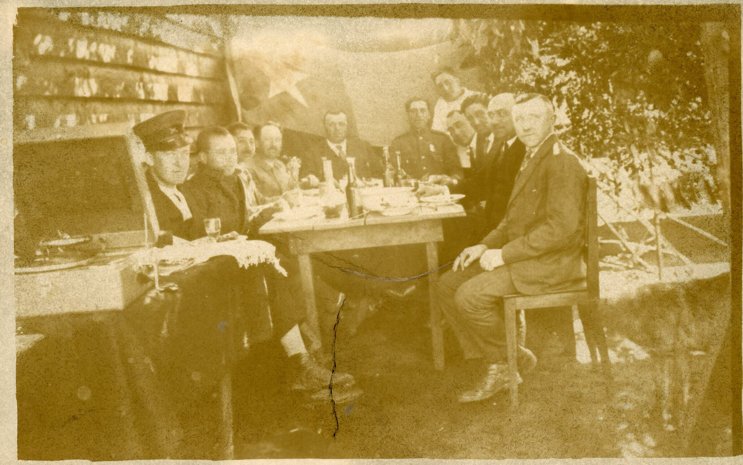 Retrato grupal de plano entero de hombres y dos Carabineros posando sentados alrededor de una mesa. En el fondo se puede observar una bandera chilena y al costado de la mesa un toca disco. Se encuentran en el patio de una casa