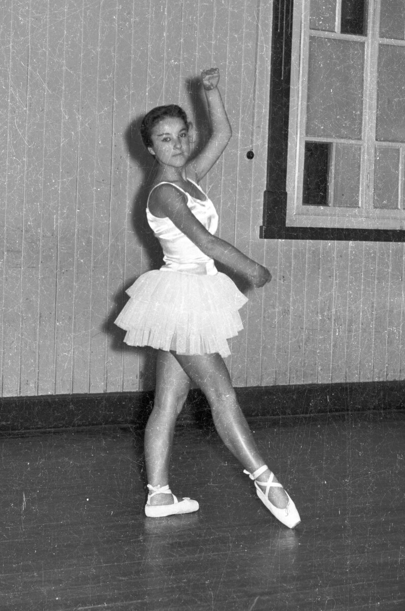 Retrato de plano entero de una mujer joven caracterizada como bailarina de ballet. está posando de pie y de Medio perfil mientras levanta las manos. Se encuentra en en el interior de un lugar no identificado