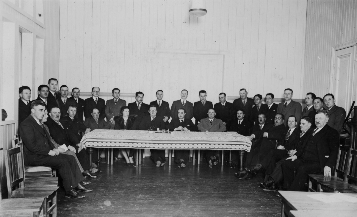 Retrato grupal de plano entero de un grupo numeroso de alcaldes de la región de Los Ríos y Los Lagos posando detrás de una mesa. Todos visten formalmente. Algunos están sentados y el resto están parados por detrás. La sexta persona sentada de izquierda a derecha es Olga Boettcher. La fotografía fue tomada durante la convención de alcaldes en la ciudad de La Unión