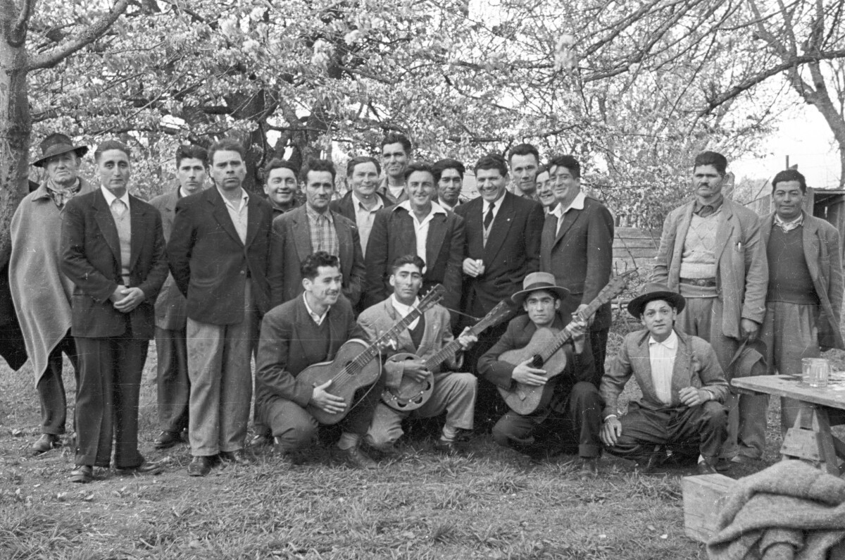 Retrato grupal de plano entero de Hombres posando formados en dos hileras. En la fila de abajo, tres hombres posando con sus guitarras. Se encuentran al aire libre en un sector no identificado