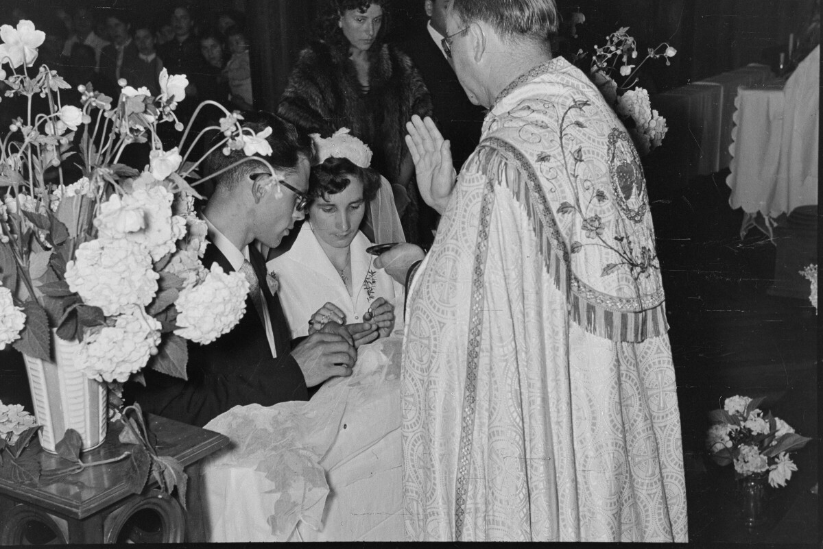 Plano entero de un sacerdote bendiciendo a una pareja de novios, quienes se encuentran hincados frente a él, en el altar. El sacerdote tiene una bandeja en la mano y la novia está colocando la argolla en la mano de su marido. Se encuentran en el interior de la parroquia San José lugar donde se está llevando a cabo la misa de celebración de su casamiento