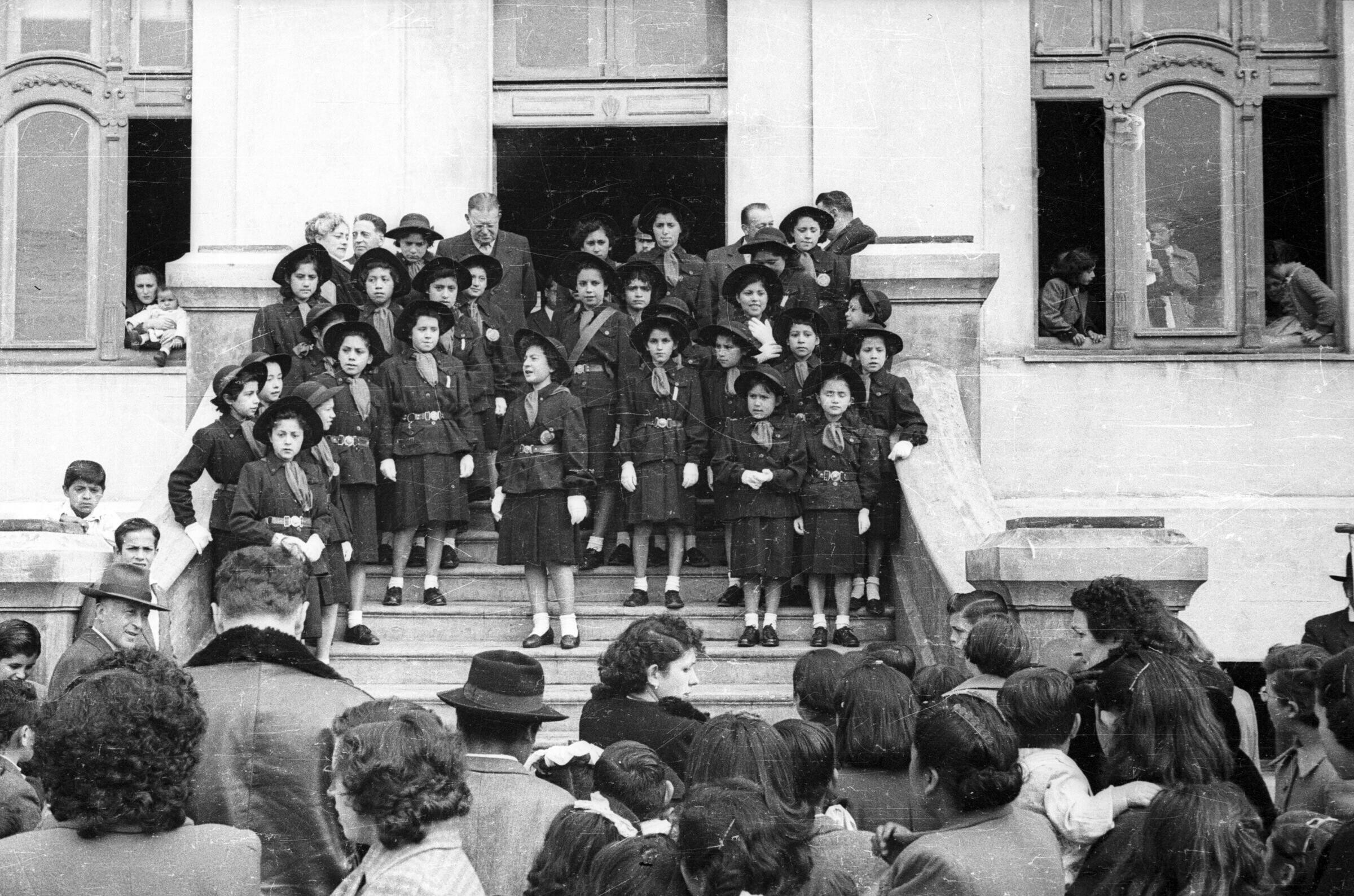 plano general de un grupo de niñas pertenecientes al grupo de scout paradas sobre unas escaleras. Una de ellas se encuentra hablándole a Mujeres, Hombres, Niños y Niñas que las observan en las afueras de la Municipalidad de La Unión.