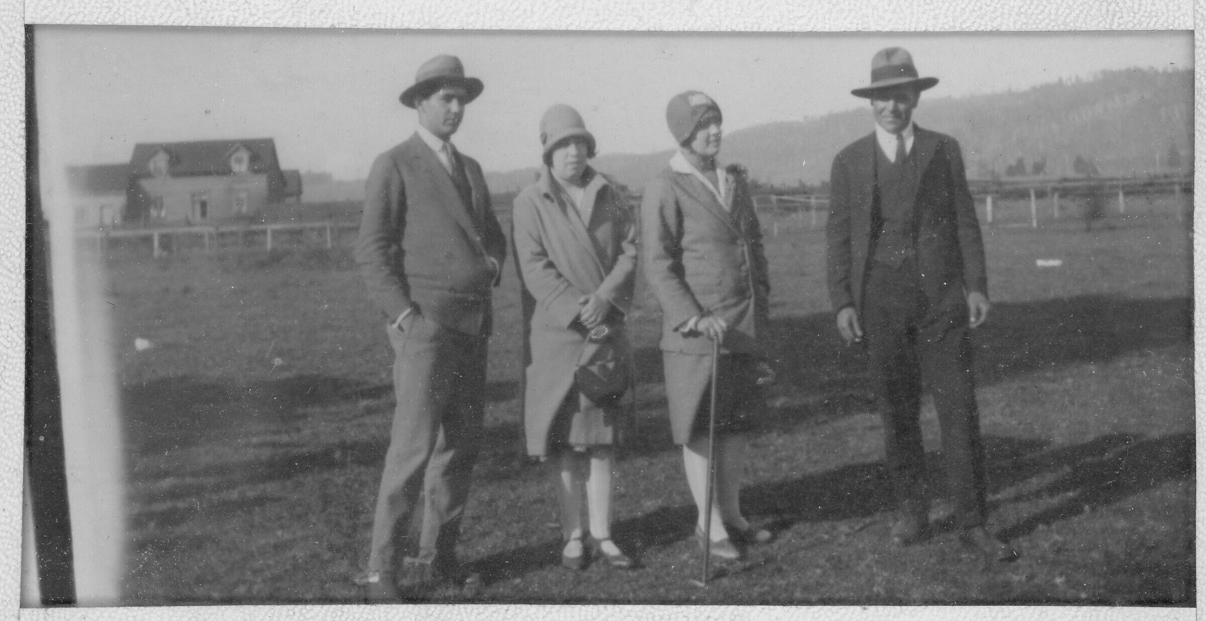Retrato de plano entero de  dos hombres y dos mujeres posando. A sus espaldas hay una casa de dos pisos y un cerro poblado de árboles. Lugar no identificado.