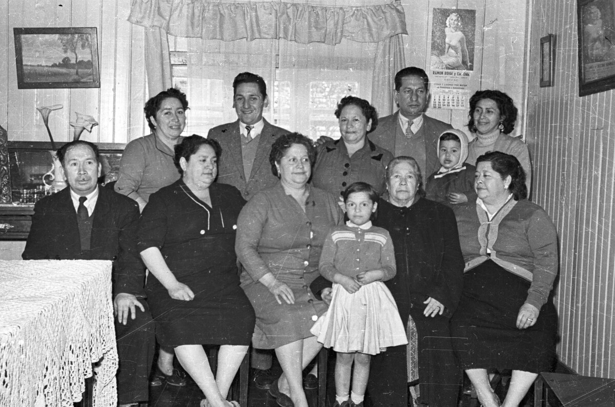 Retrato grupal de plano entero de Mujeres, Hombres, un bebé y una niña posando formados junto a una ventana en el interior de una casa