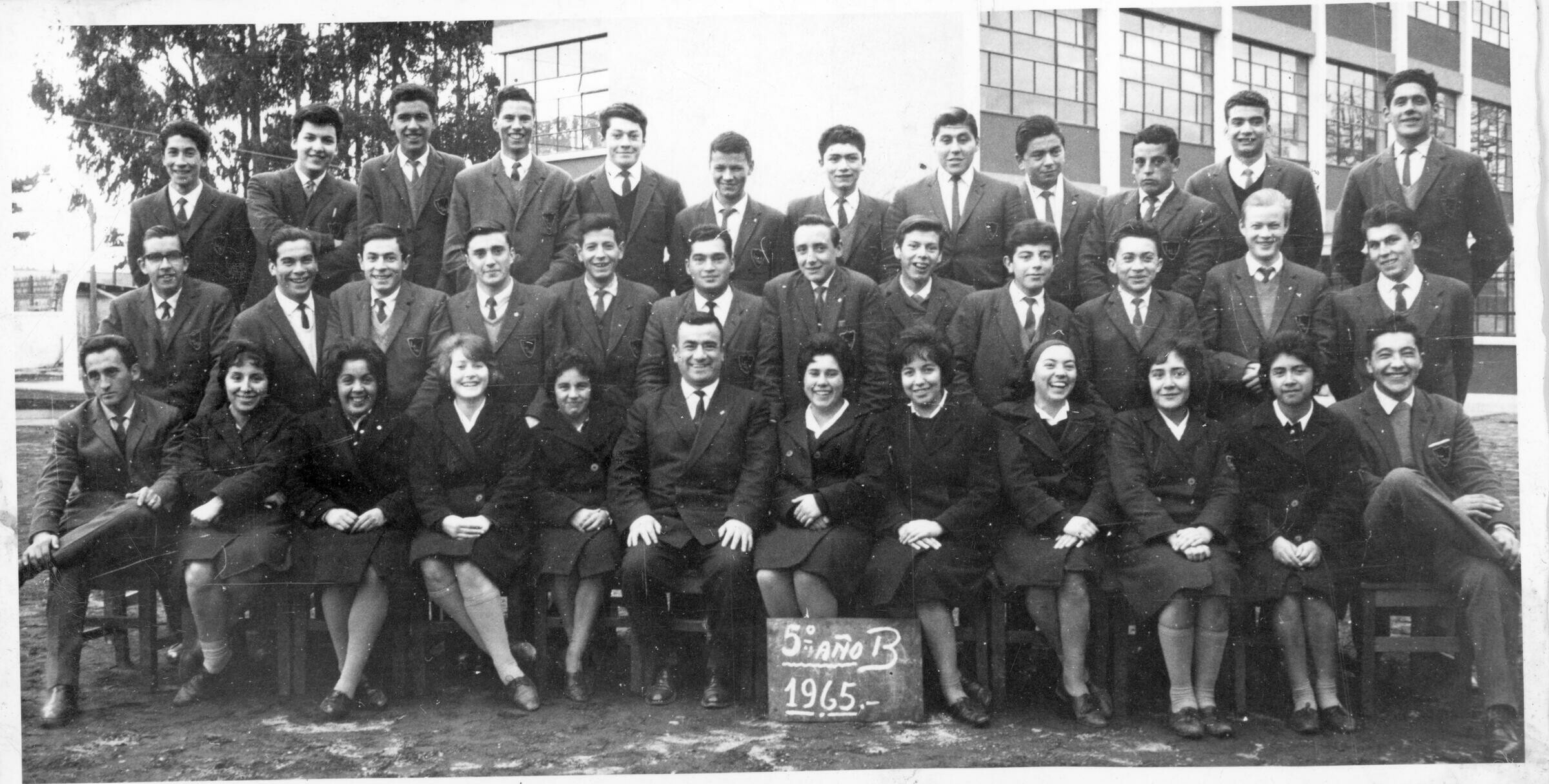 -Retrato grupal de plano entero de Mujeres y Hombres, formados junto a un profesor, quien se encuentra posando en primera fila. Todos sonríen mientras posan. En el suelo, apoyado sobre las piernas de una de las estudiantes hay un letrero que dice --5º año B 1965.-- Se encuentran en el patio del liceo La Unión-