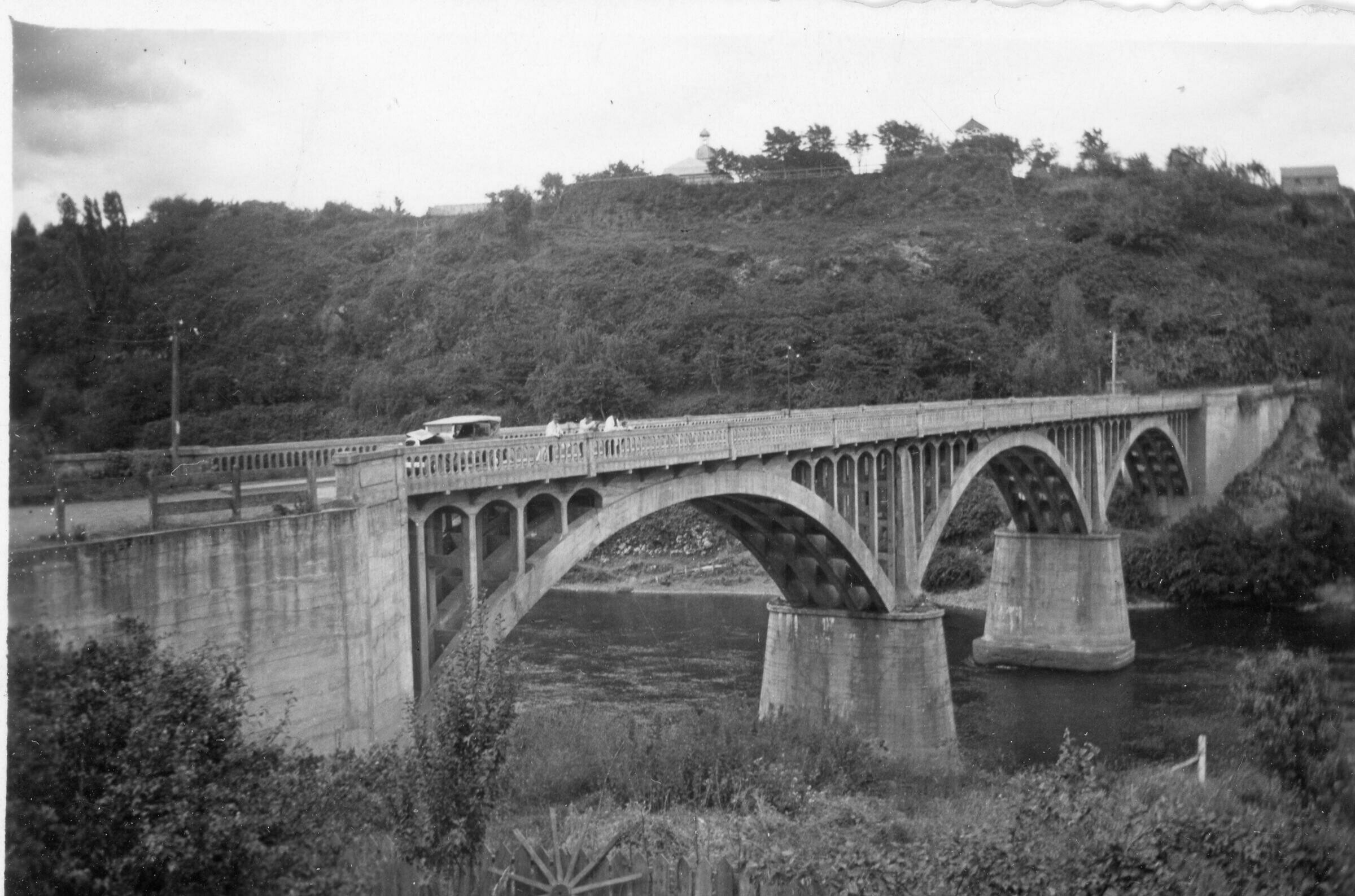 Plano general de tres hombres mirando hacia el rio bueno desde la baranda del  puente Carlos Ibáñez del Campo. A sus espaldas va pasando un automóvil.