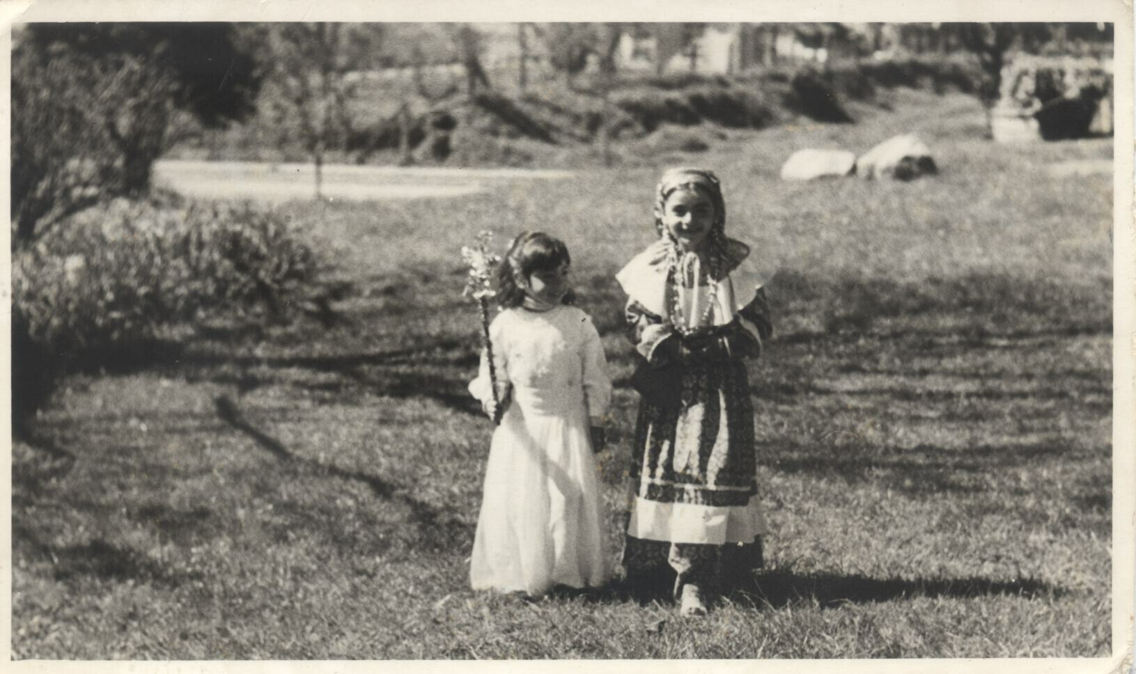 Plano entero de dos niñas disfrazadas. A la izquierda una está disfrazada de hada y a la derecha está disfrazada de gitana. Se encuentran paradas sobre el pasto, en un sector abierto no identificado.