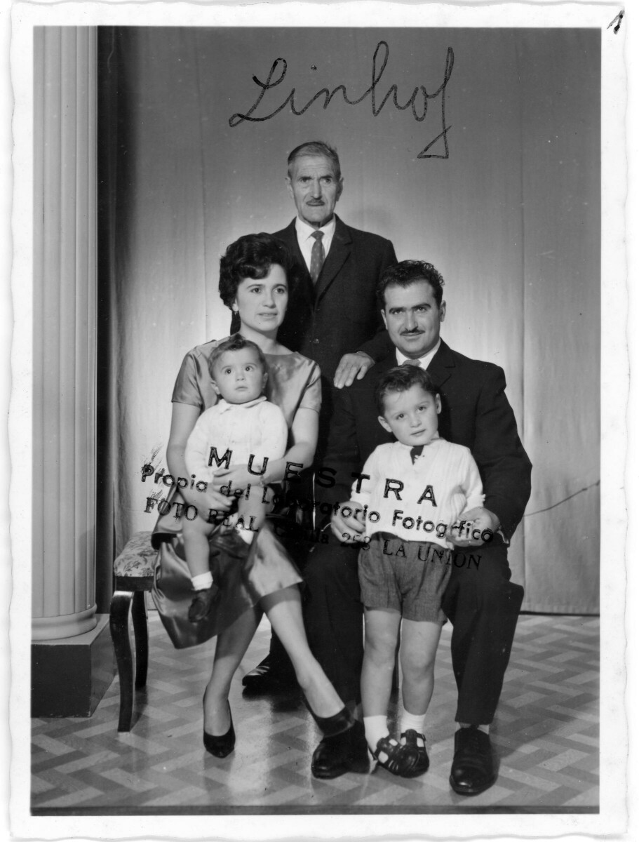 Retrato de estudio Plano entero de una mujer posando sentada con un niño sobre sus piernas. En la silla de al lado, hay un hombre de Traje, posando con un niño parado entre sus piernas y por detrás de ambas sillas, hay un hombre mayor, vestido de Traje, posando parado