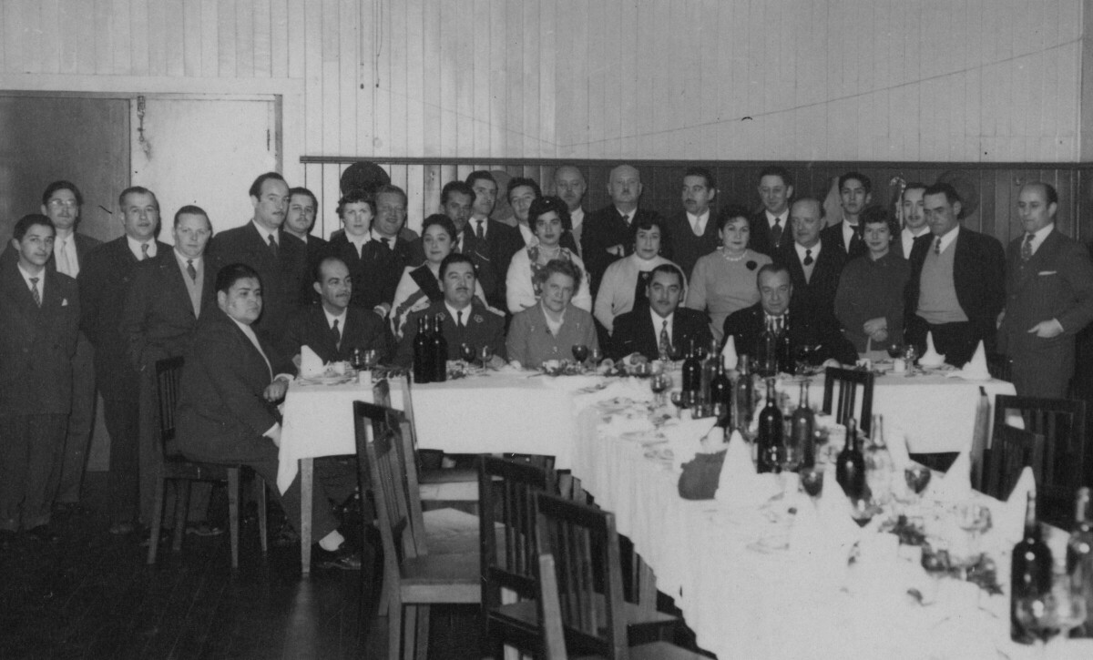 Retrato grupal de plano entero de Hombres, Mujeres, un carabinero y Olga Boettcher, posando de frente detrás de una mesa. Olga y el uniformado se encuentran sentados en el centro de la primera fila. Se encuentran en el interior de una infraestructura no identificada durante una celebración