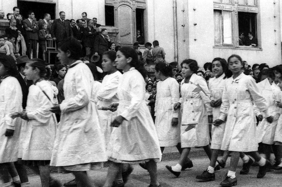 plano general de un grupo de niñas con delantal, desfilando formadas en hileras. En el fondo, un grupo de personas y Carlos Vogel, se encuentran observándolas en el frontis de la municipalidad