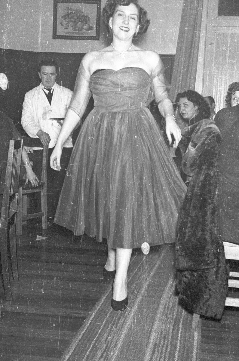Plano entero de una mujer modelando un vestido. Está parada entre un grupo de personas que se encuentran sentadas alrededor de unas mesas. En el fondo hay un garzón parado con una bandeja en la mano. Está en el interior de un local mientras se realiza una exhibición de Vestidos