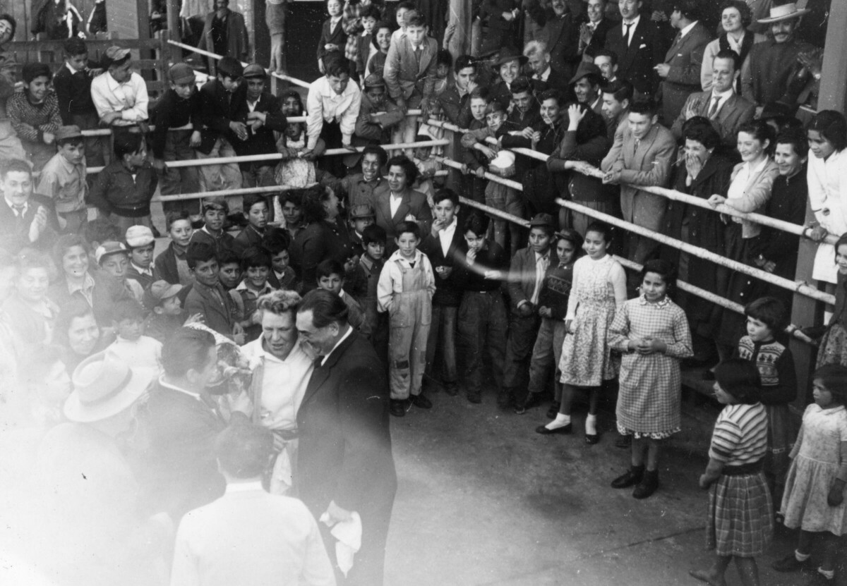 plano general de Olga Boettcher abrazada con su acompañante de baile, mientras un hombre les entrega un ramo de flores una vez finalizado el pie de cueca. A su alrededor, un grupo numeroso de Niñas, Niños, Hombres y Mujeres los acompañan en el interior de un recinto no identificado