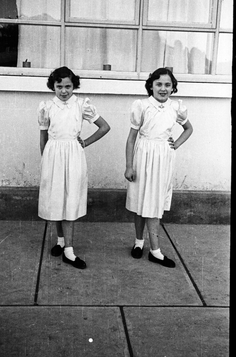 Retrato de plano entero de dos niñas posando, ambas llevan puesto un vestido blanco y zapatos negros. Están en el patio de un colegio