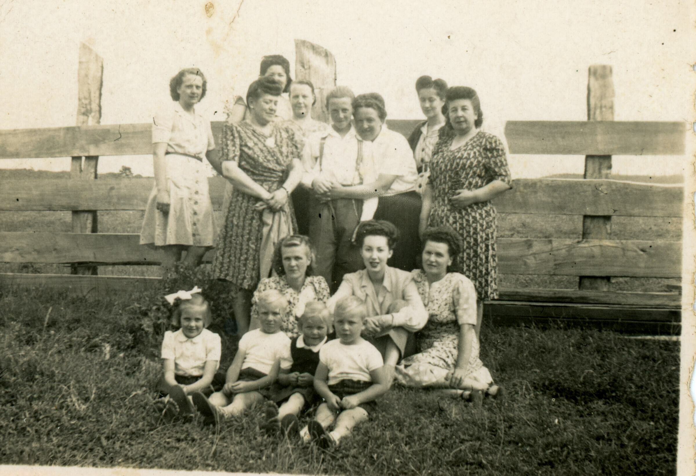 Retrato grupal de plano entero de mujeres, niñas y niños posando delante de un cerco de madera en un lugar no identificado.