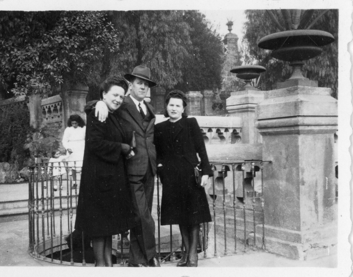 Retrato de plano entero de dos mujeres y un hombre posando apoyados en una reja metálica. A sus espaldas, una niña camina de la mano de una mujer. Lugar no identificado.