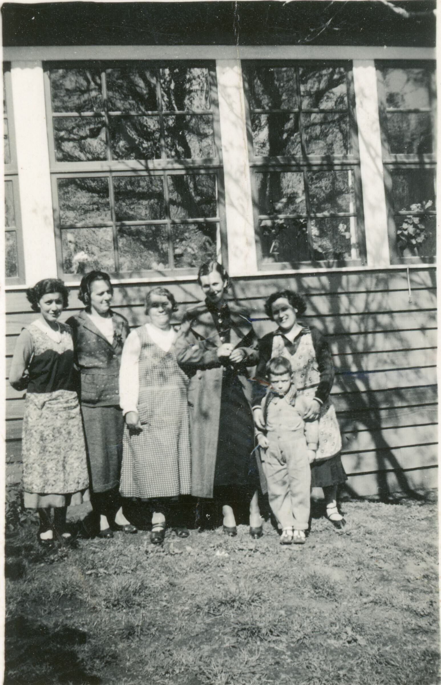 Retrato grupal de plano entero de mujeres y un niño posando junto a una casa con ventanales. Lugar no identificado.