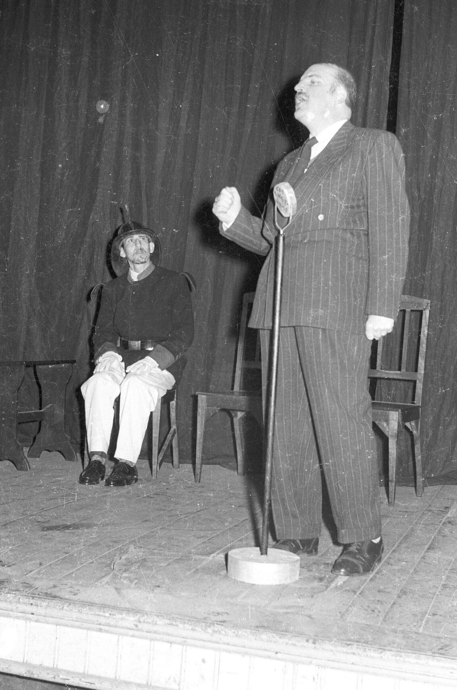 Plano entero de un hombre de Traje hablando por un micrófono mientras a sus espaldas un bombero sentado lo observa. Ambos están representando una obra de teatro en el interior del gimnasio de la escuela superior de niñas Nº2