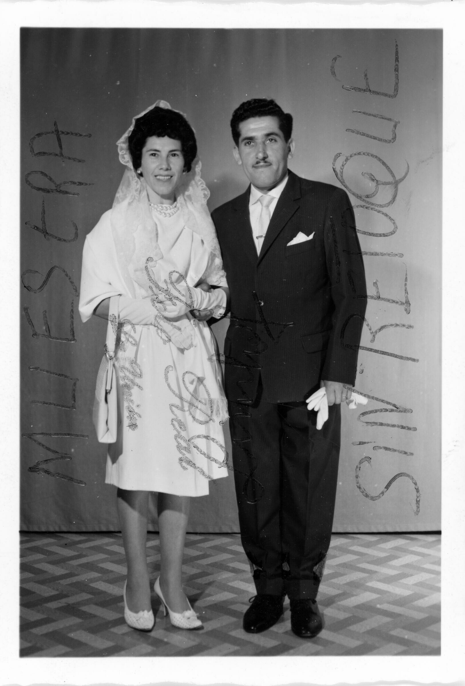 Retrato de estudio de Plano entero de Omar Peralta y su novia, posando tomados del brazo. La novia lleva colgando una cartera y el novio tiene en la mano sus guantes. Ambos se encuentran parados sonriendo