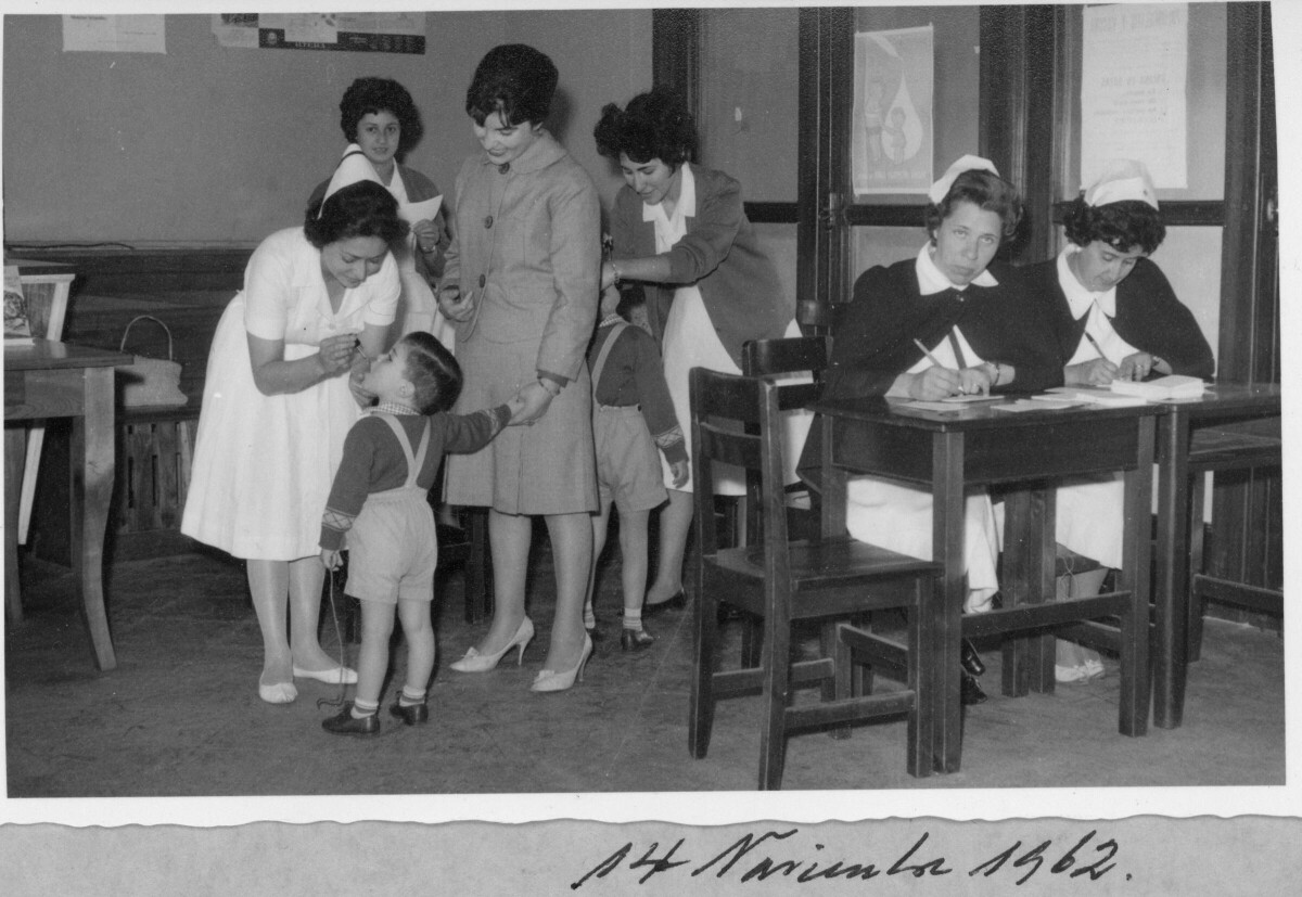 plano general de una enfermera poniendo gotitas en la boca de un niño mientras este toma la mano de una mujer. Dos enfermeras se encuentran sentadas mientras una de ellas está mirando hacia la cámara. Otro Niño está parado en el fondo mientras una enfermera le toma la cara. Se encuentran realizando exámenes de rutina en el interior de una infraestructura no identificada.