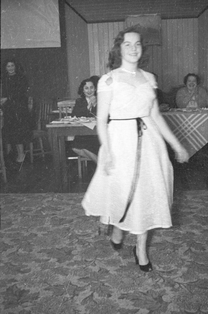 Plano entero de una mujer modelando un vestido, en el fondo hay un grupo de mujeres que la aplaude mientras la observan sentadas alrededor de unas mesas en el interior de un local