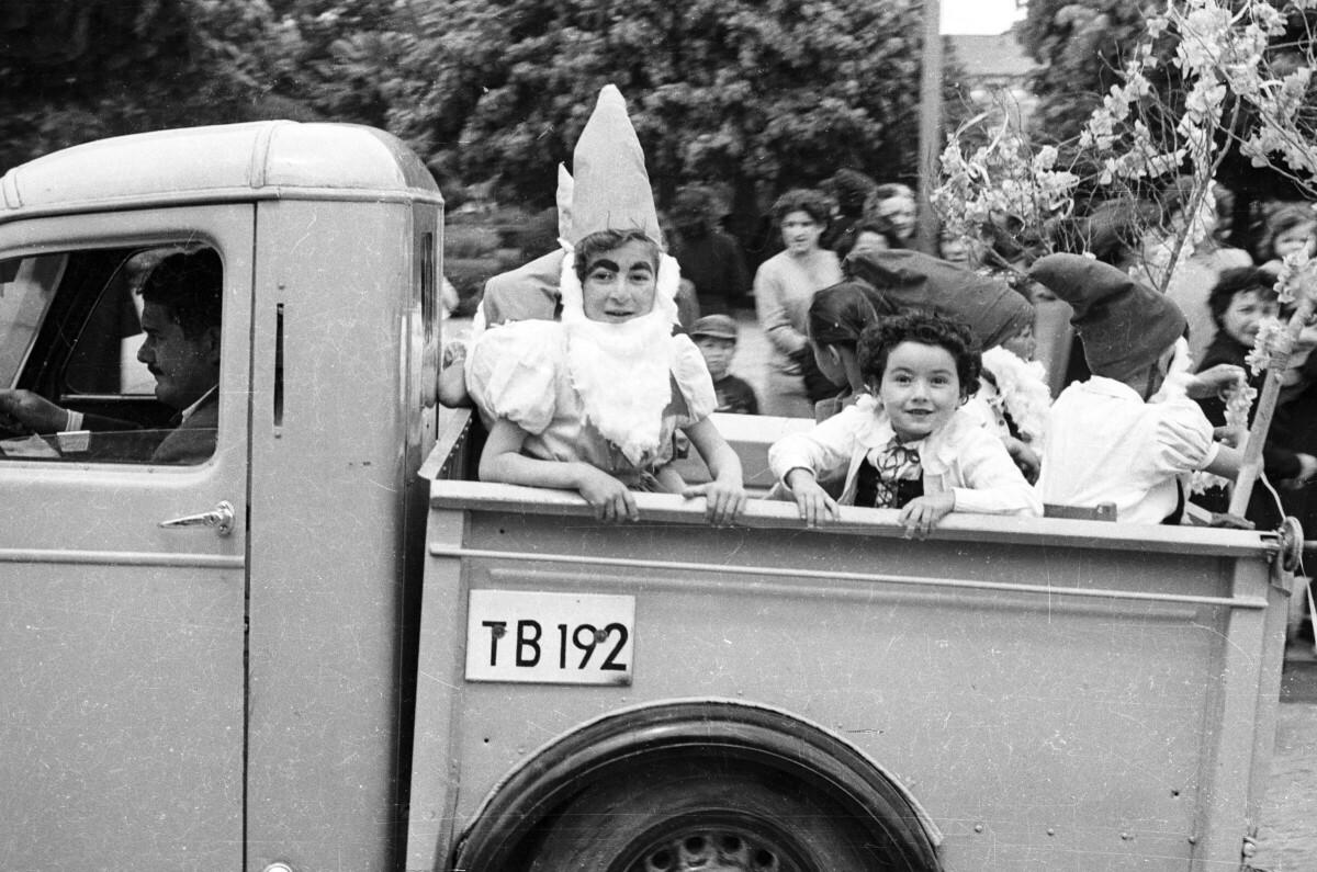 Plano entero de un niña y un grupo de Niños disfrazados de blanca nieves y enanitos, mirando hacia la cámara desde la parte trasera de una camioneta la cual es conducida por un hombre vestido de Traje. Van avanzando en un desfile de carros alegóricos por un costado de la Plaza de La Concordia