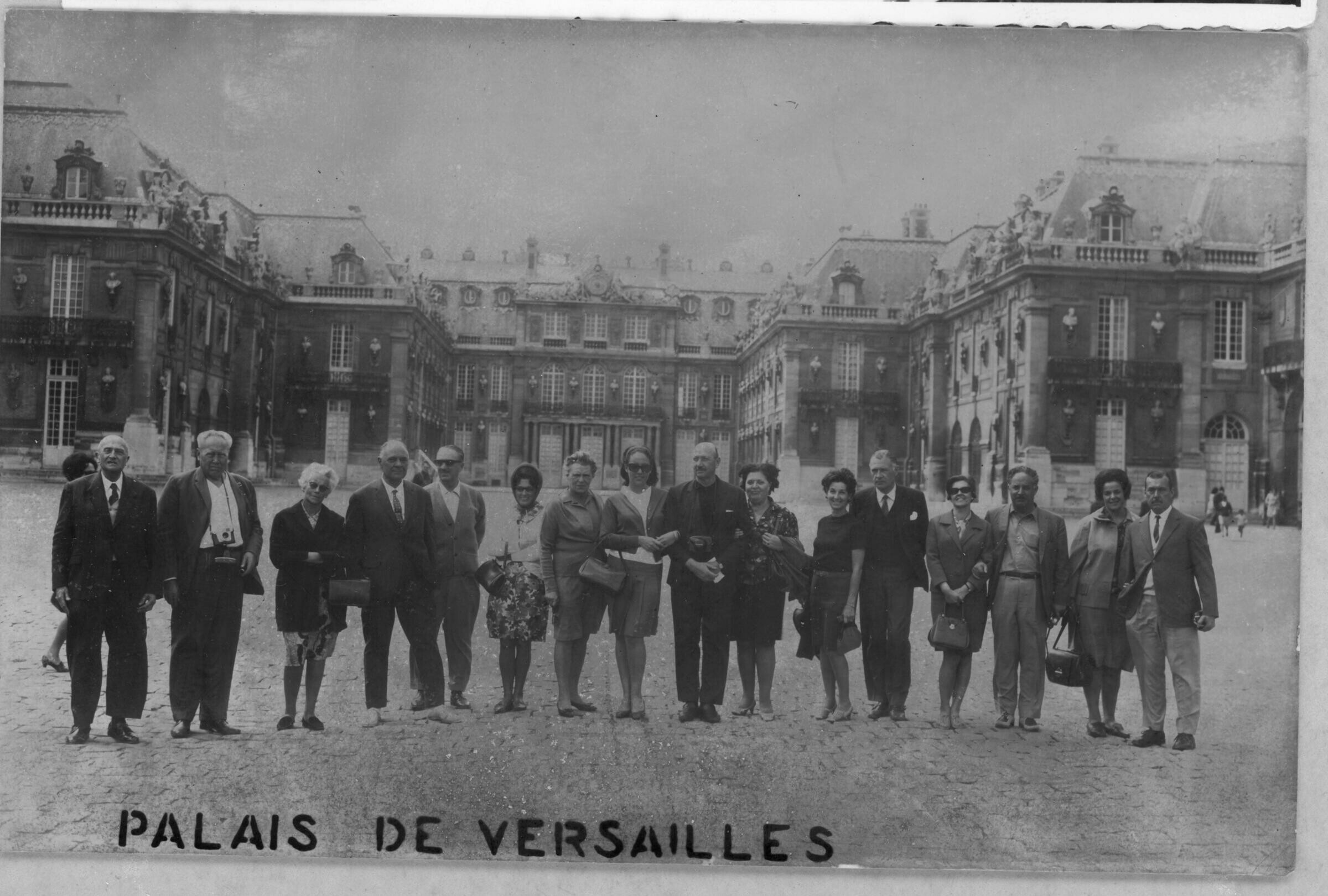 Retrato grupal de plano entero de Hombres y Mujeres posando en un hilera. Algunas de las mujeres llevan sus carteras y dos de los hombres tienen una cámara colgando desde el pecho. En el fondo de la imagen se ve el palacio de Versalles, lugar al que visitan durante un viaje a Francia.