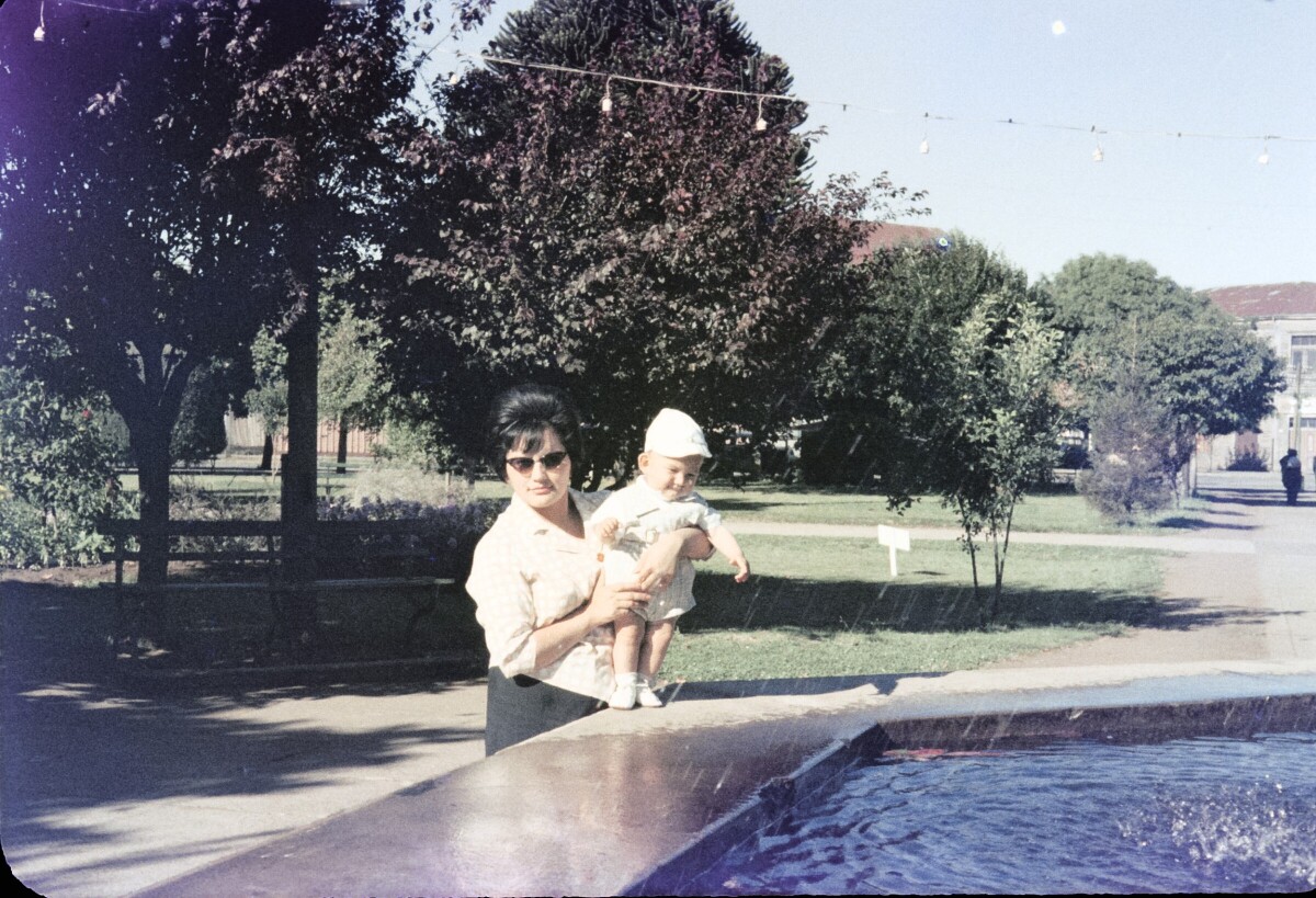Retrato de plano medio de mujer con vestido con un bebé en brazos, sosteniéndolo de pie sobre la pileta de agua en plaza de la Concordia de La Unión