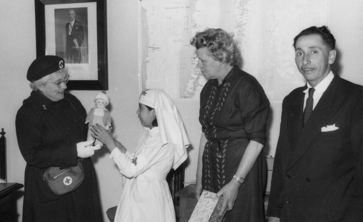 Plano entero de una mujer mayor perteneciente a la cruz roja, recibiendo una muñeca que le entrega una Niña, voluntaria de la misma institución. A su lado, las acompaña Olga Boettcher quien también es miembro y un hombre vestido de Traje. En la pared del fondo hay un retrato presidencial y mapas pegados en la pared. Se encuentran en el interior de una infraestructura no identificada.