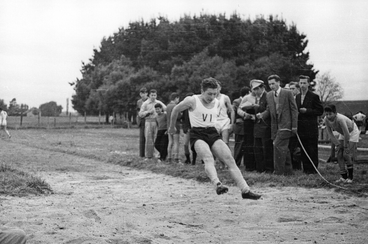 Plano entero de un hombre aterrizando en la pista tras haber realizado un salto largo, en un costado hay un grupo de Hombres y Niños observándolo durante las competencias de juegos olímpicos en un recinto abierto