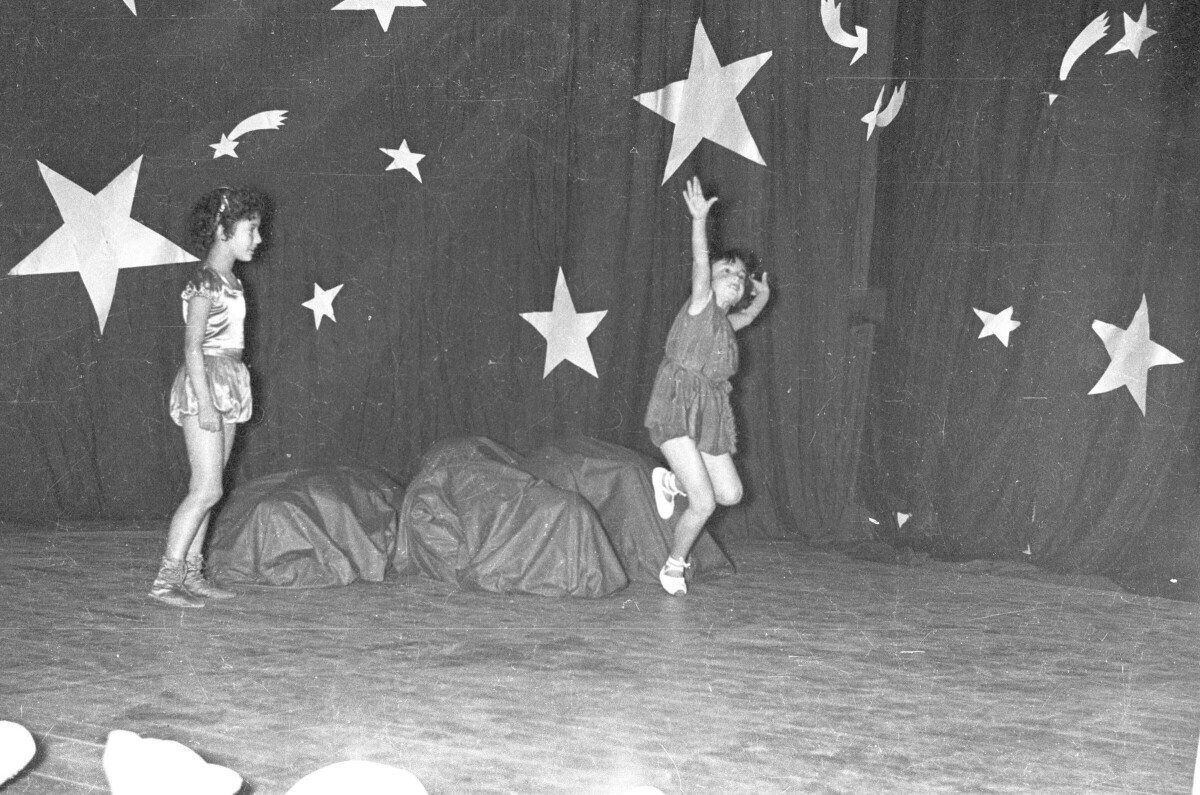 Plano entero de dos niñas bailando ballet durante la presentación de una obra de teatro. En el fondo el telón está decorado con estrellas de utilería. Están en el interior de una escuela durante una jornada de presentaciones artísticas.