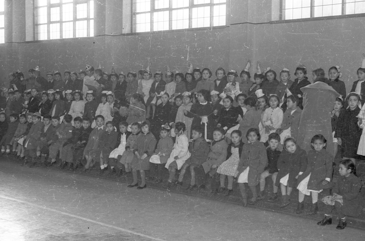 plano general de gran cantidad de niñas y Niños, algunos con gorros de cumpleaños, sentados en las galerías del gimnasio de Escuela superior de niñas n°2