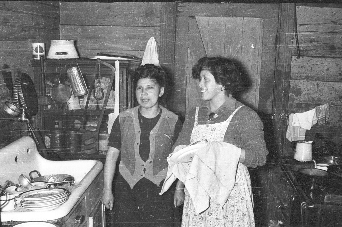 Plano entero de dos mujeres de pie junto a un lavaplatos. Una de las mujeres lleva puesto un delantal mientras seca un plato con un mantel. En el lavaplatos hay varia loza esperando por secar tales como platos, un cucharon y tazas. Están en el interior de la cocina de una casa