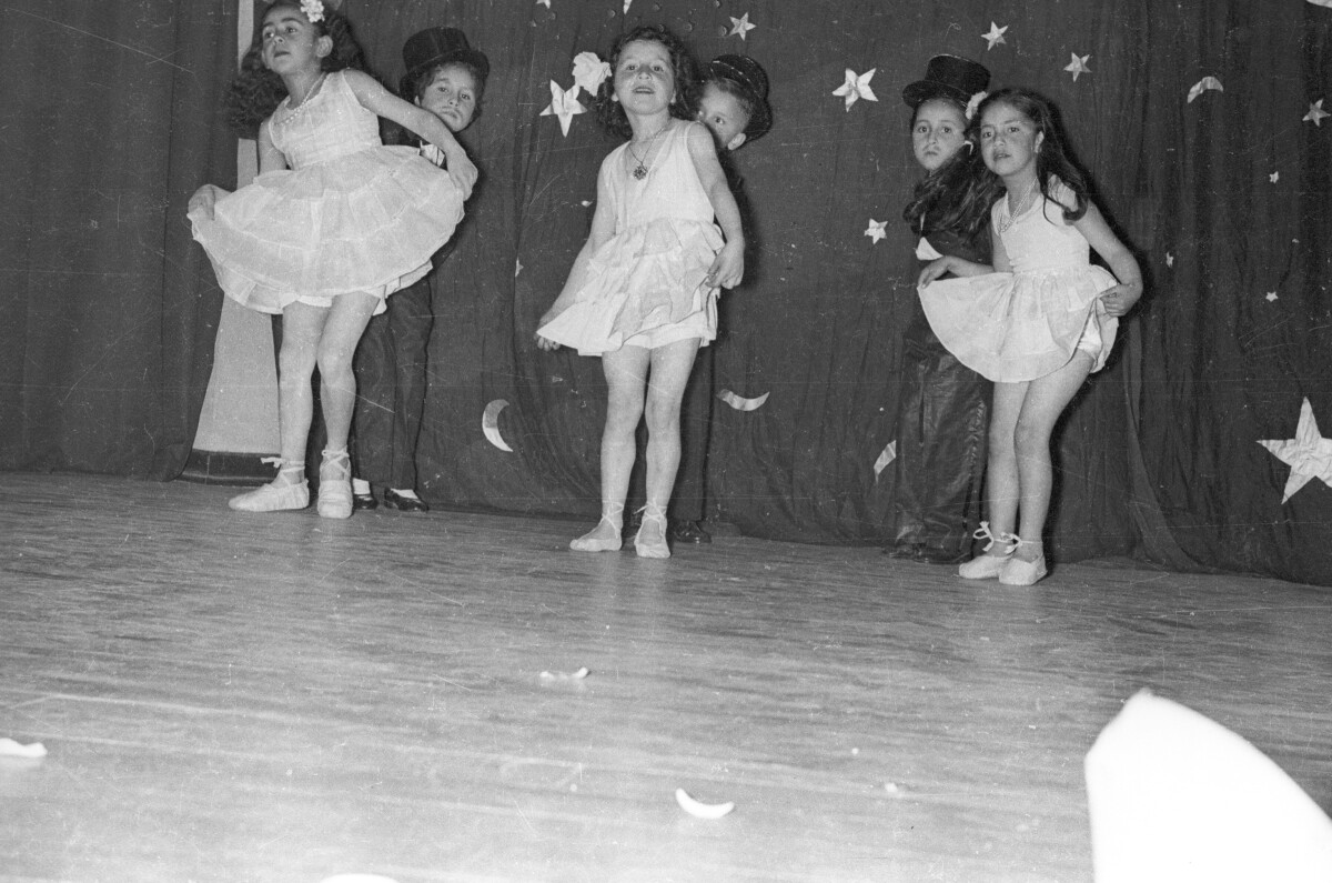 Plano entero de tres niñas y tres niños bailando en parejas sobre un escenario el cual tiene un telón en el fondo decorado por estrellas de utilería. Están realizando la presentación de un número artístico en el gimnasio de la escuela super de niñas Nº3