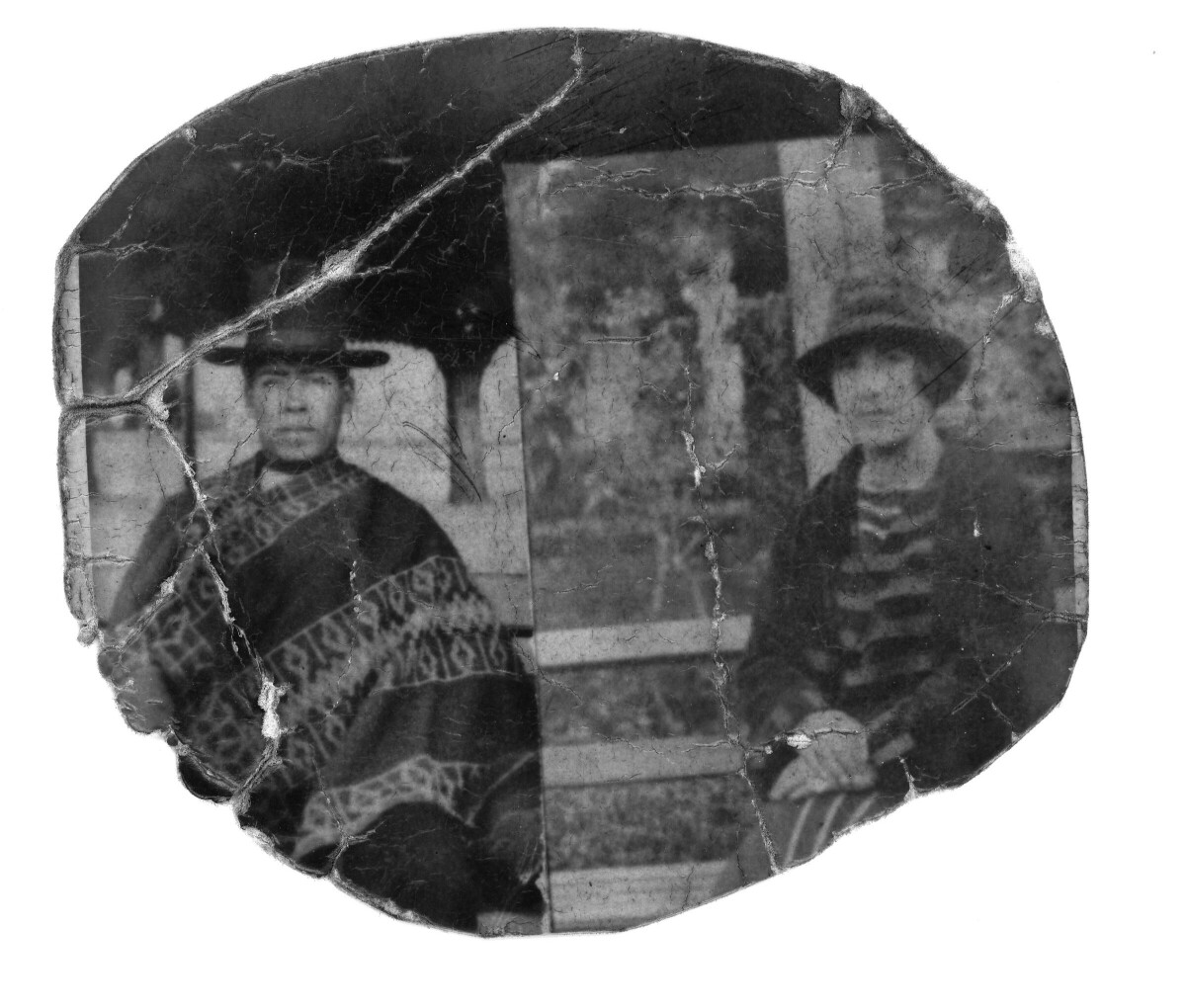 Retrato de plano medio largo  de un hombre de manta y sombrero, posando sentado junto a una mujer con sombrero en la banca de una plaza no identificada