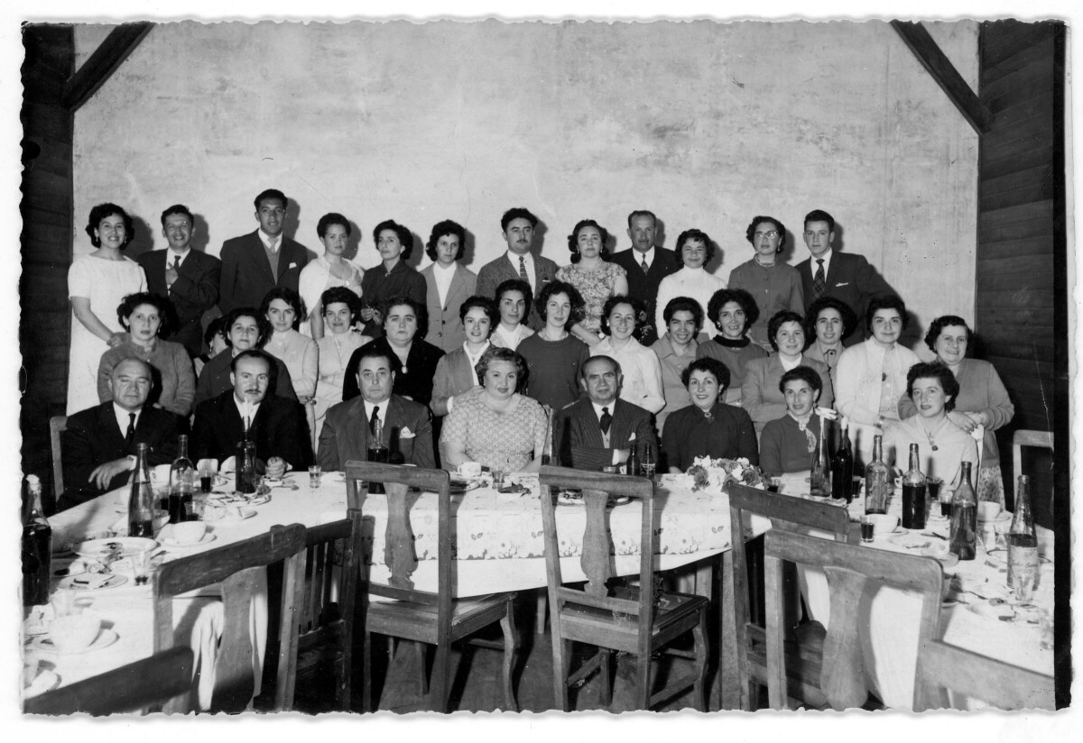 Retrato grupal de plano entero de Mujeres y Hombres posando detrás de una mesa. Dado el gran número de personas es que se encuentran formados en tres hileras, en la primera están sentados y en las otras dos están de pie. Sobre la mesa hay botellas y platos, Todos visten formalmente. Se encuentran en el interior de una infraestructura no identificada durante una celebración
