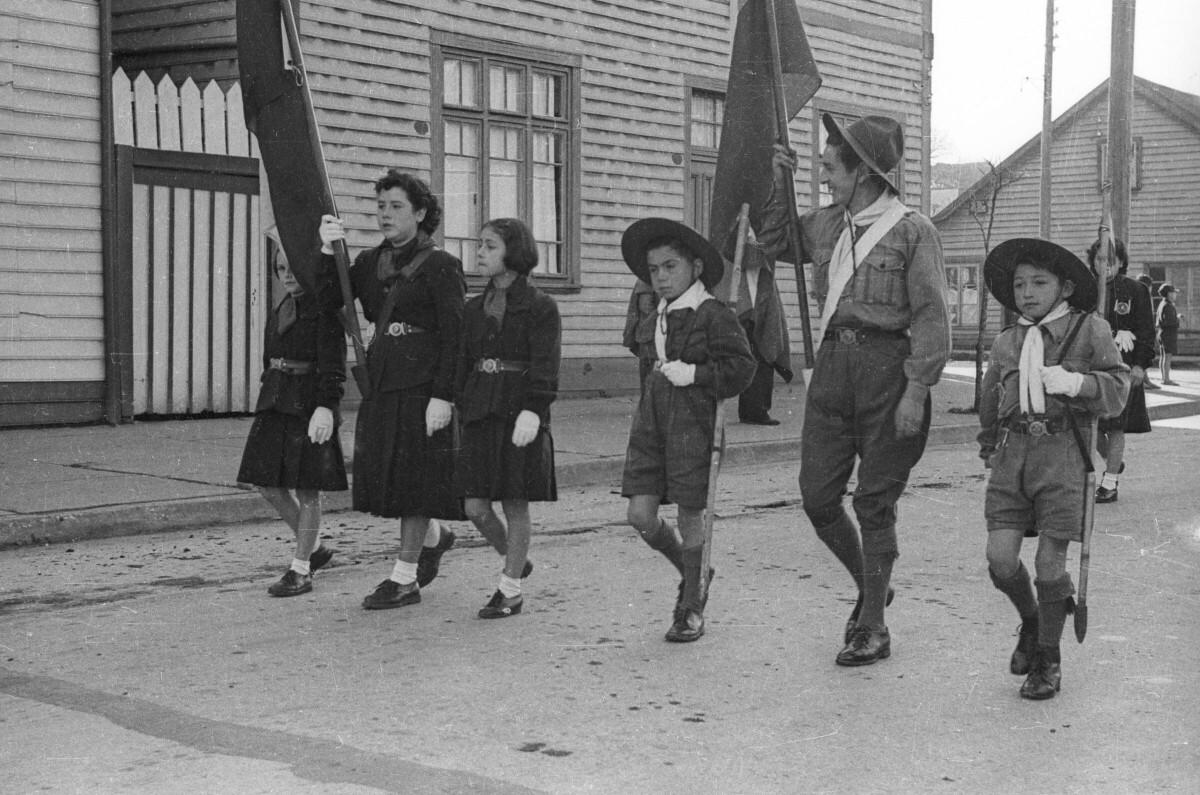 Plano entero de tres niñas y tres niños caminando formados en una filera. La Niña y el Niño de mayor edad, llevan izando una bandera tomándola por el mástil. Todos son pertenecientes a la brigada de scout y llevan puesto su uniforme. Van desfilando por una de las calles de la ciudad de La Unión