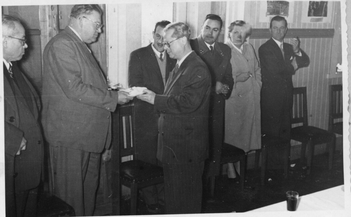 Plano entero de dos hombres tomando un plato. Ambos visten de Traje y usan lentes. En el fondo, un hombre, un carabinero y Olga Boettcher los están observando mientras tiene un cigarro en la mano. Están ubicados detrás de una mesa, en el interior de una infraestructura no identificada, durante un banquete