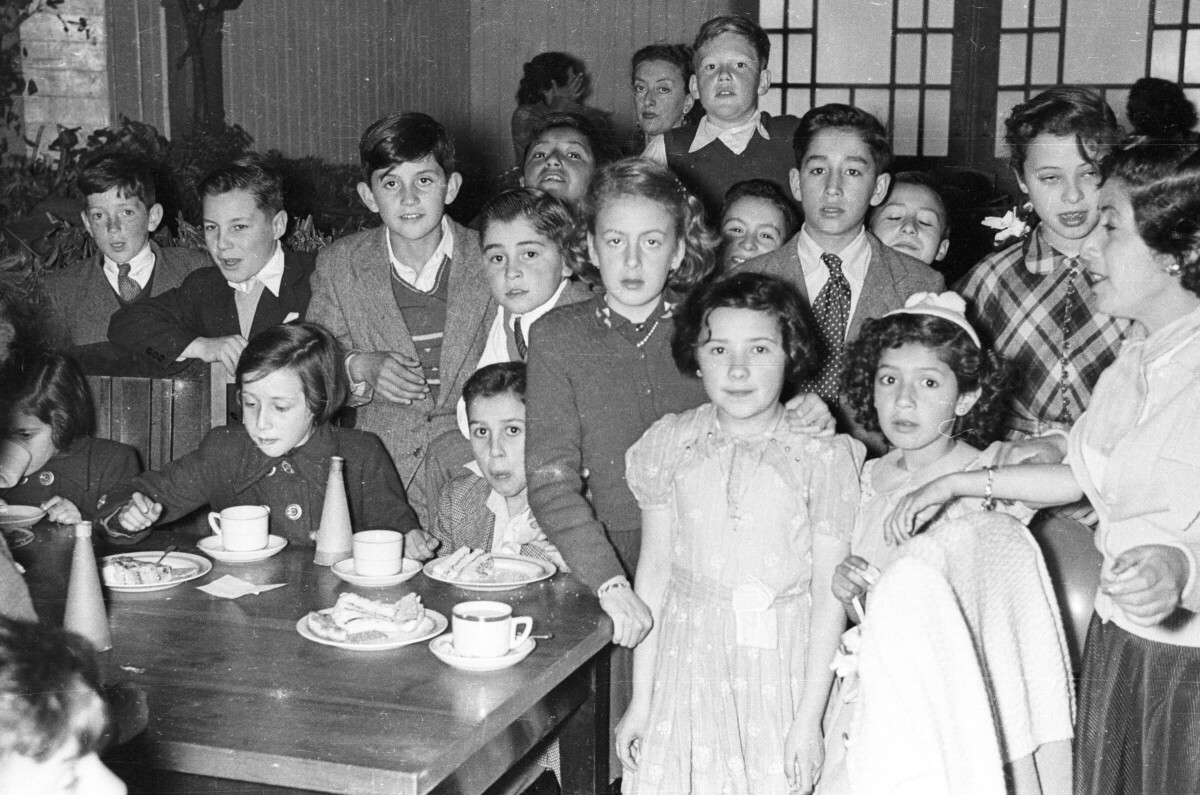plano general de un grupo de Niños y Niñas parados detrás de una mesa. Algunos se encuentra mirando hacia la cámara, otros están sentados junto a la mesa comiendo y otros se encuentran mirando hacia otros lugares. Junto a ellos hay una mujer parada que les está hablando. Están en el interior de un local no identificado durante la celebración de fiestas patrias.