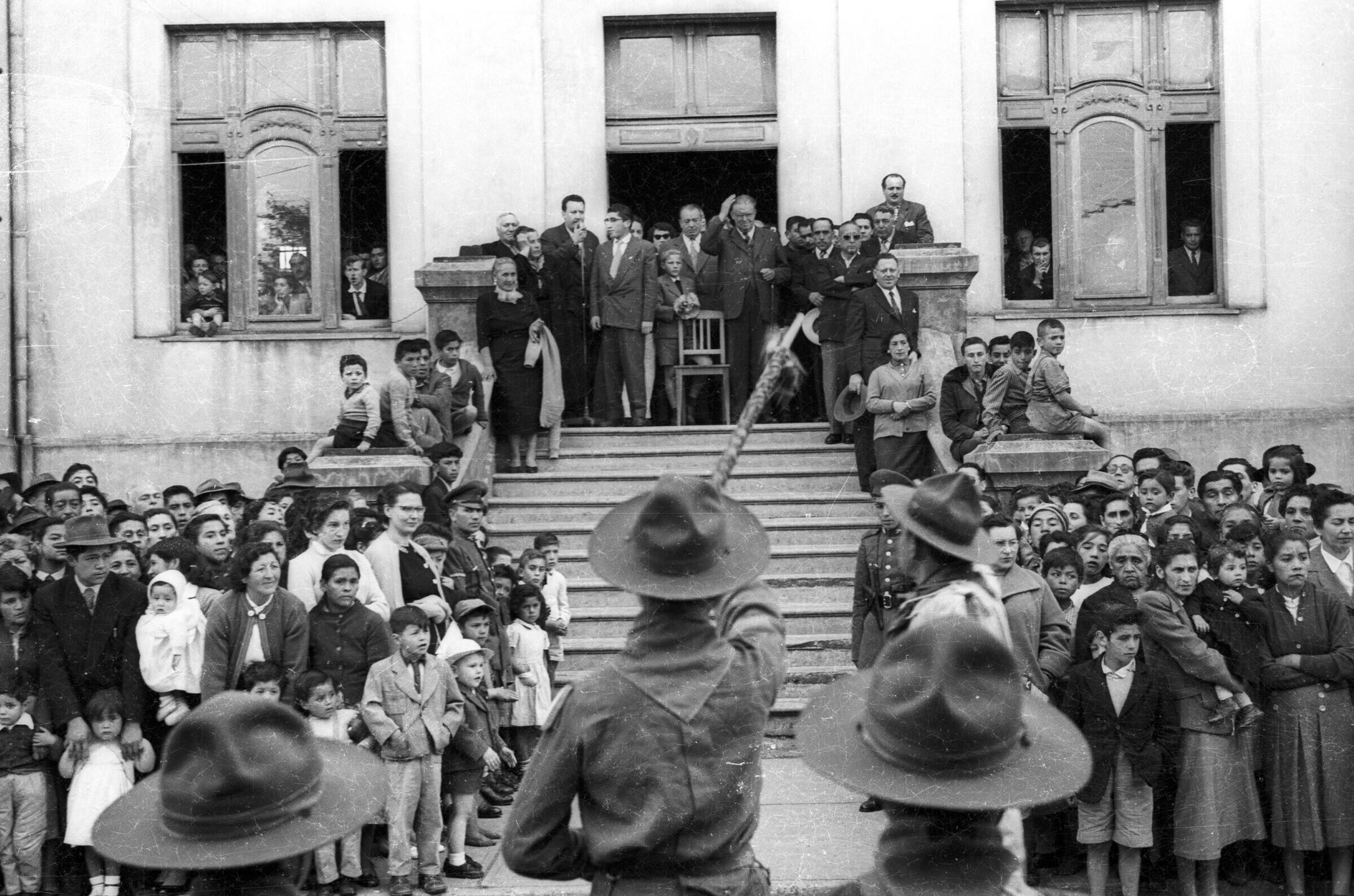 Plano entero de un grupo de niños de una brigada no identificada, realizando un homenaje frente a Carlos Vogel, quien se encuentra en el fondo saludandoles desde el frontis de la municipalidad