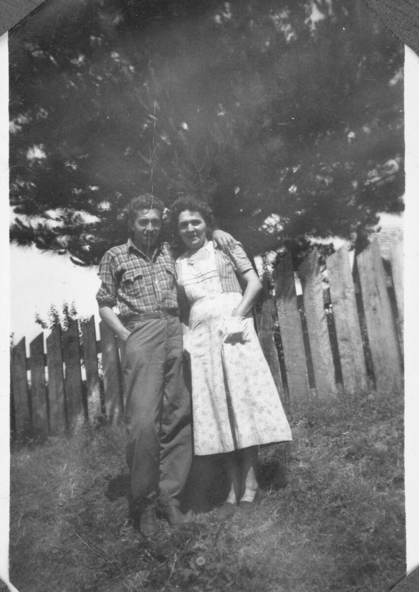 Retrato de hombre y mujer adultos en entorno rural. El hombre viste camisa y pantalón de tela, y la mujer una falda y blusa. detrás hay un cerco de madera y un árbol.