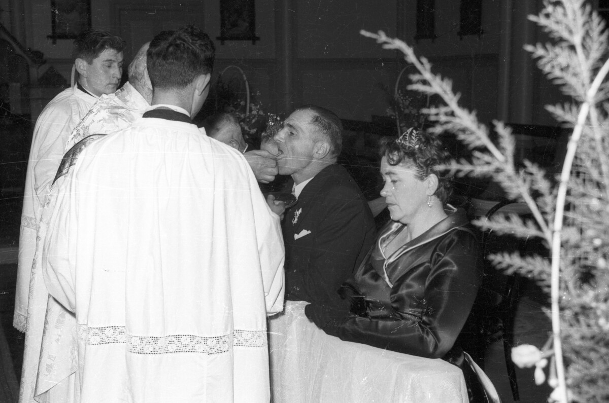 Plano medio de Mujer con Vestido y Hombre con Trajes arrodillados en reclinatorio, frente a ellos un sacerdote con 2 acólitos dando la hostia a la Mujer. En una iglesia no identificada