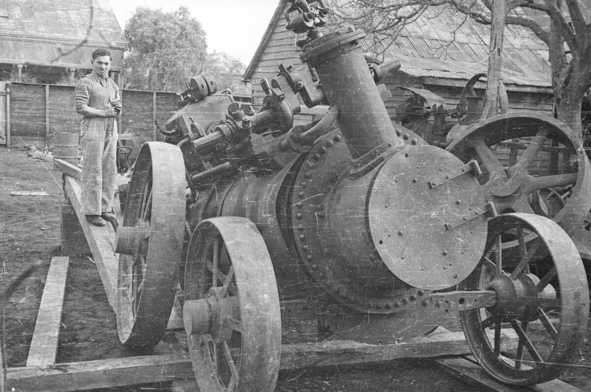 Plano entero de una maquina llamada locomóvil en mantención, en un costado sobre una viga hay un hombre de pie con una llave francesa. La maquina está en el patio de una casa